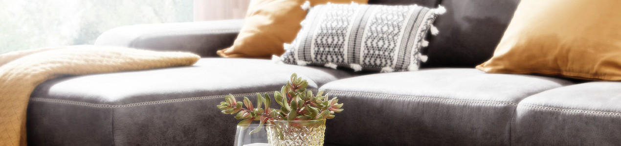 Graues Sofa mit gelben und gemusterten Kissen, davor Glas mit kleiner Pflanze auf dem Tisch.