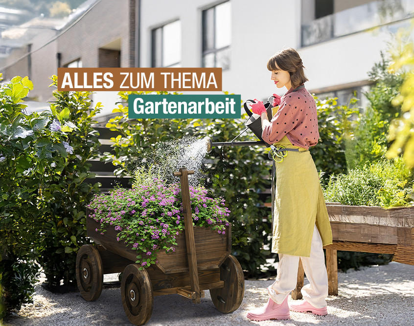 Person mit gelber Schürze gießt Pflanzen in einem hölzernen Gartenwagen vor modernen Wohngebäuden