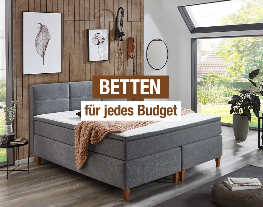 "BETTEN für jedes Budget". Graues Boxspringbett im modernen Schlafzimmer mit Holzdeko und viel Tageslicht.