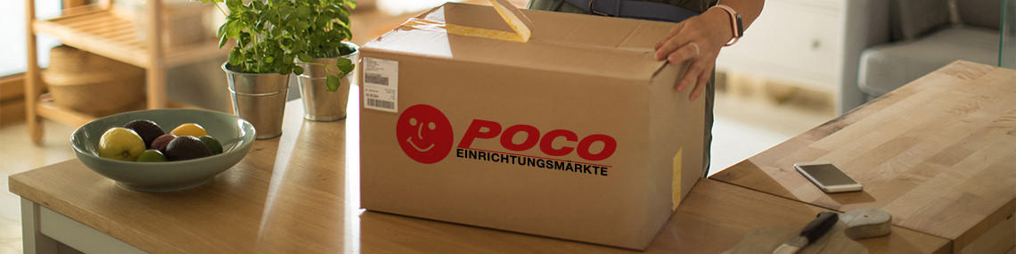 Person packt ein Paket von POCO aus auf Holztisch neben Obstschale und Smartphone