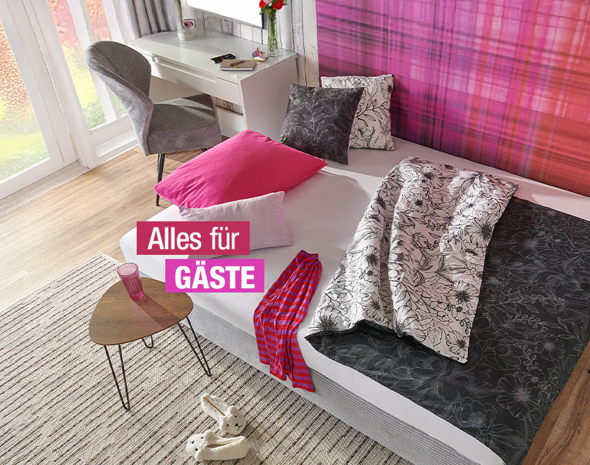 Gästezimmer mit Einzelbett, pinkem Kissen, gemustertem Bettbezug, Schreibtisch mit Stuhl und Fenster mit Vorhängen